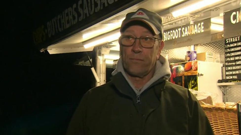 A man wearing a coat, jumper and cap looks at the camera as he stands in front of a food van. 