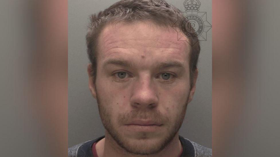 A police custody shot of Alex Fullard looking at the camera. He has short brown hair light brown facial stubble. He is wearing a grey top with a black neckline. The wall behind him is grey.