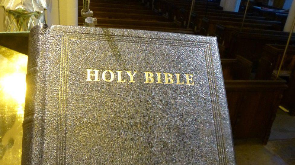 A leather bound bible with the words 'Holy Bible' in capital letters, printed in a gold coloured typeface on its front. Wooden pews inside St Peter's Church can be seen behind. 
