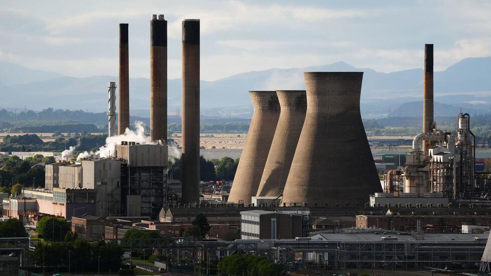 An oil refinery with tall buildings emitting smoke. Mountains are seen in the background.