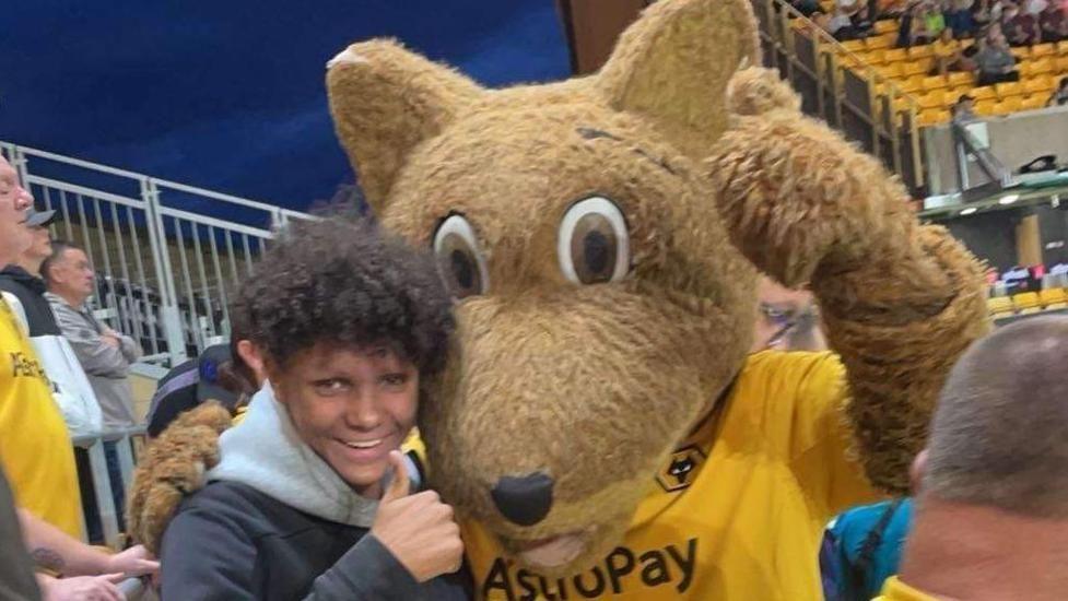 Harleigh Hepworth, a Wolverhampton Wanderers fan, is seen with a Wolves mascot at a game. He has his arm around the mascot, with one thumb up, and a big smile. He is wearing a grey hoodie with a black top.