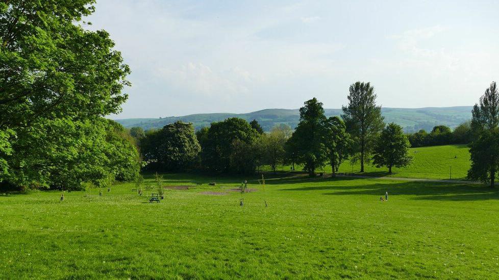 A large green space with several shrubs and trees. Rolling hills can be seen in the distance on what is a sunny day.