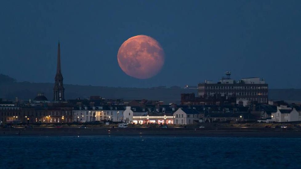 The image shows a large, full, orange moon in the night sky above Ayr town centre. There is a large body of water in the foreground. The townscape is silhouetted against the moon.