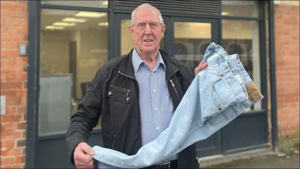 Elderly man in a shirt and dark blue coat holds up a pair of denim jeans