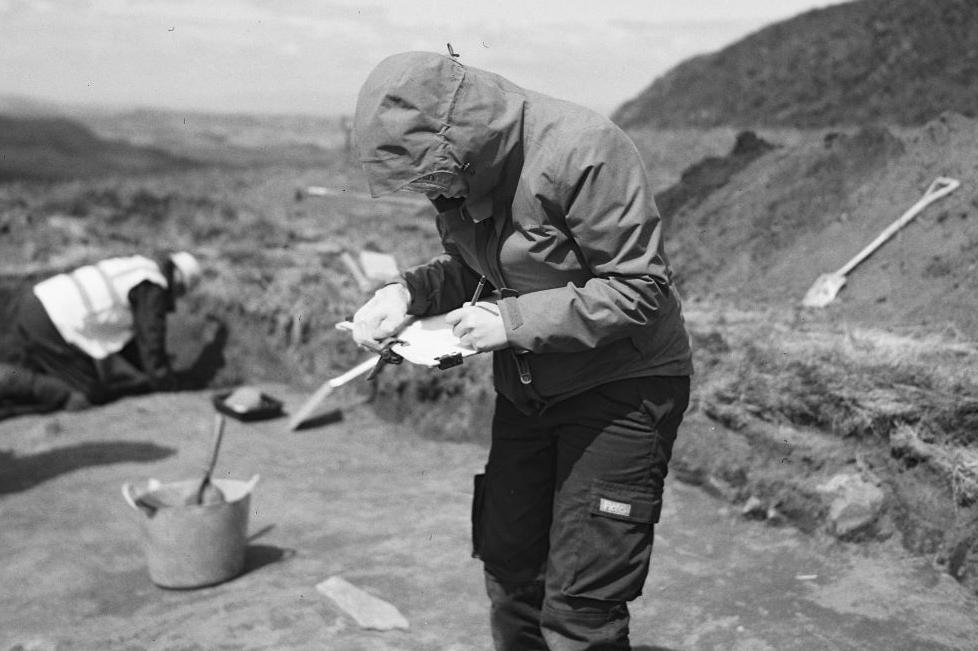 An archaeologist, with the hood of their jacket up over their head, writing notes