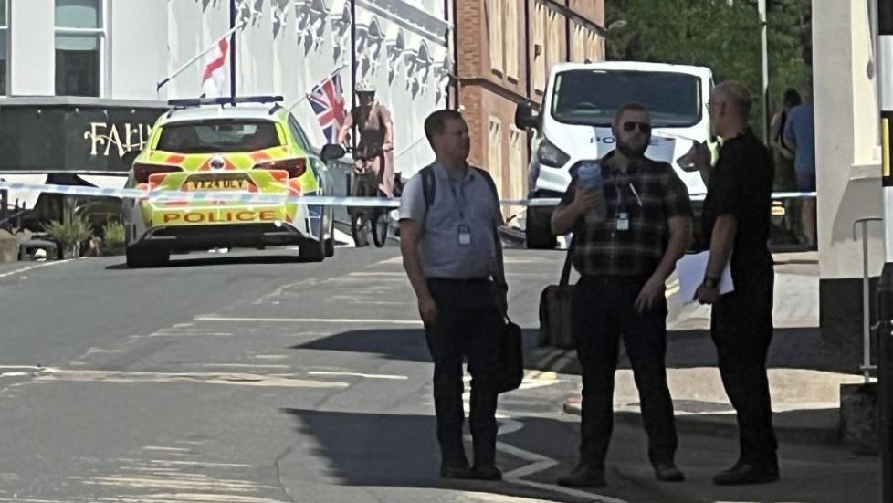 Three men standing in an empty street in shadow with a police car and a row of white buildings in the background as well as a police car