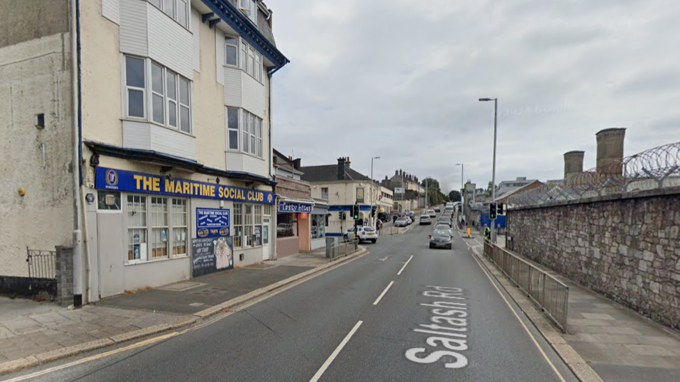 Google street view of The Maritime Social Club on Saltash Road in Plymouth. The four-storey building has two bay windows on the mid two floors, below there is a blue sign with yellow writing saying 'The Maritime Social Club' and other signs with writing that cannot be made out. Parked and driving cars are visible in the background. Overcast sky.