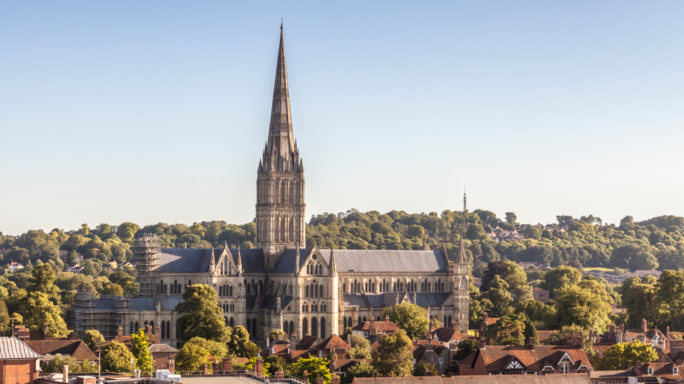 A wide shot of the Salisbury landscape including the cathedral with green trees surrounding it. There is a blue sky in the background.