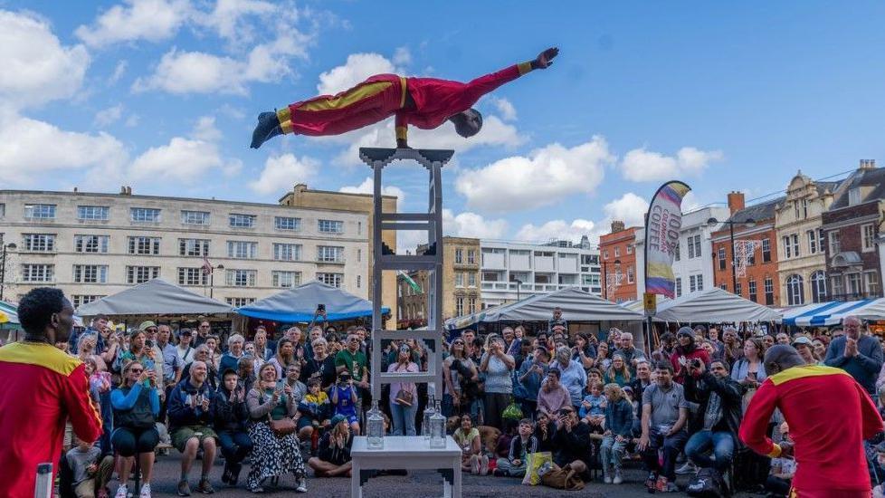 A man is balancing on one arm on a ladder or plinth high up above crowds of people gathered in Market Square in Cambridge. He is wearing a red and yellow outfit and there are two similarly dressed men on the ground underneath him. The people in the crowd are smiling and clapping. Old ornate brick and stone buildings can be seen in the background