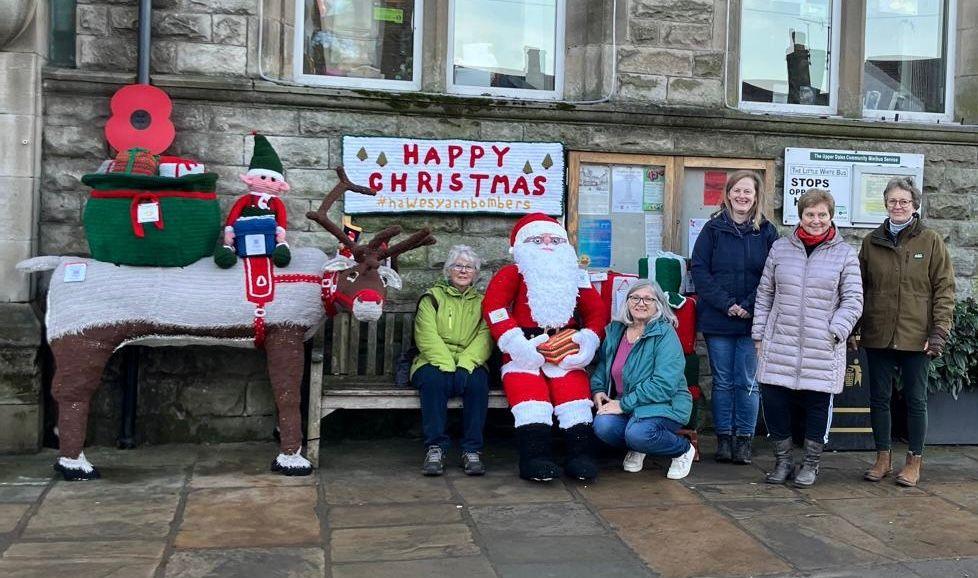 A knitted reindeer is pictured next to a bench on which sits one of the knitters and a smaller, knitted Santa, while the other knitters crouch and stand nearby