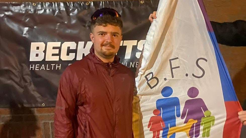 A man standing outside in front of a black banner reading "BECKETTS HEALTH AND WELLNESS" in white bold writing. He is wearing a red waterproof zip-up jacket and has sunglasses on his head. He has brown hair and dark facial hair. A hand is just in shot, holding up a colourful banner with "B.F.S" written on it as well as artwork of a family with two children and a dog.