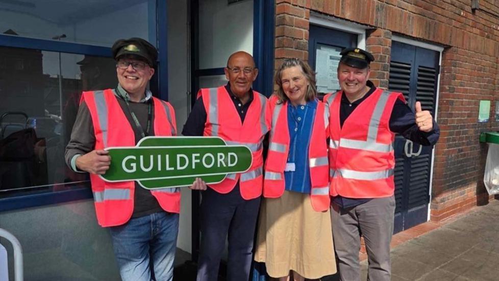 Train enthusiasts pose for a photograph while wearing bright pink, high-vis vests. A man on the left wearing a railway worker's cap holds a green sign reading 'Guildford'. They are stood in front of a brick wall at Guildford railway station.