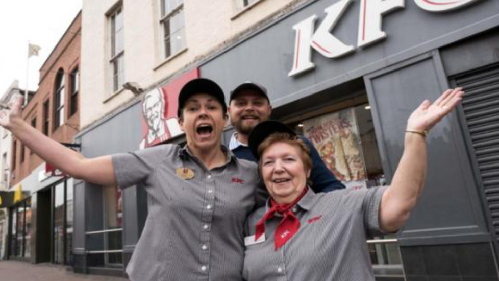 Pauline in her KFC uniform of grey shirt and red neckerchief, being hugged by two other members of the team outside a KFC. 