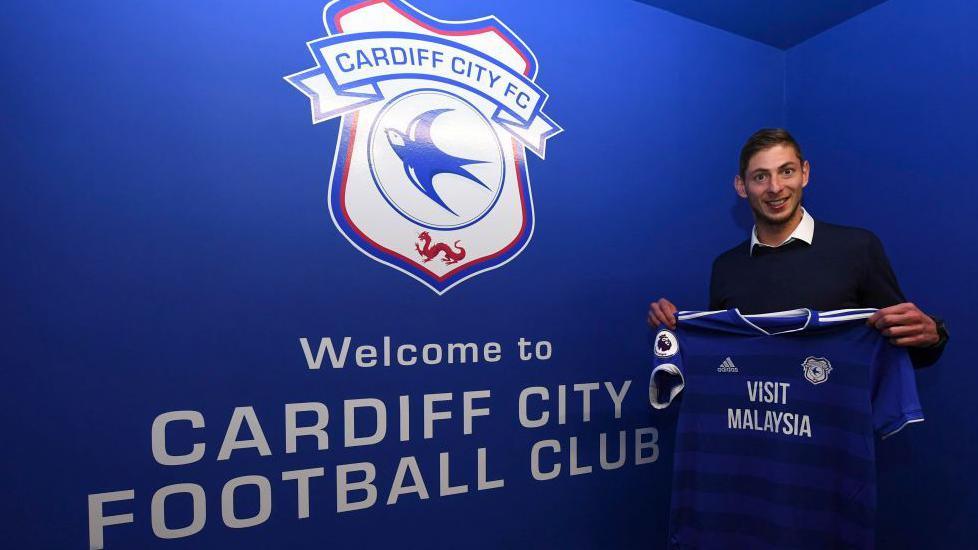 A young man with brown hair, wearing a white shirt and dark jumper. He is holding a blue Cardiff City football shirt and is stood in front of a blue, Cardiff City branded wall which reads "Welcome to Cardiff City Football Club". He is smiling and looking at the camera.