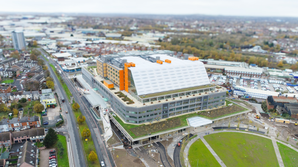 A aerial view of a new super hospital which has a white sloped roof and orange frame, with roads and a large grass area surrounding it