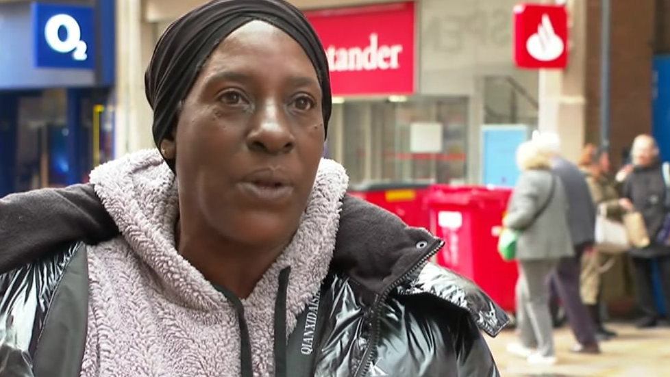 Monica is wearing a headscarf and fluffy jumper underneath a grey winter jacket. She is stood in a street in Wolverhampton.