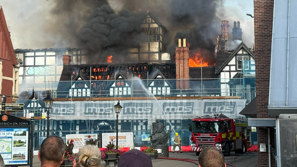 An old white building with black timber frames surrounded by scaffolding with flames and smoke coming from the roof