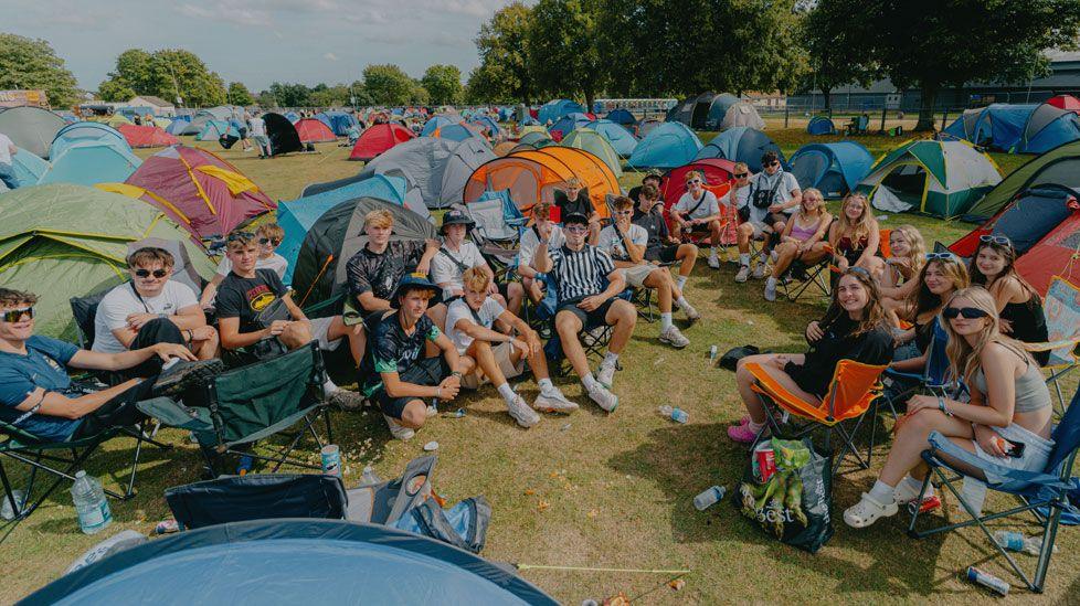 A semi-circle of about 20 young people, sitting on camping chairs or the ground, looking towards the camera and smiling with a sea of colourful pop-up tents behind them