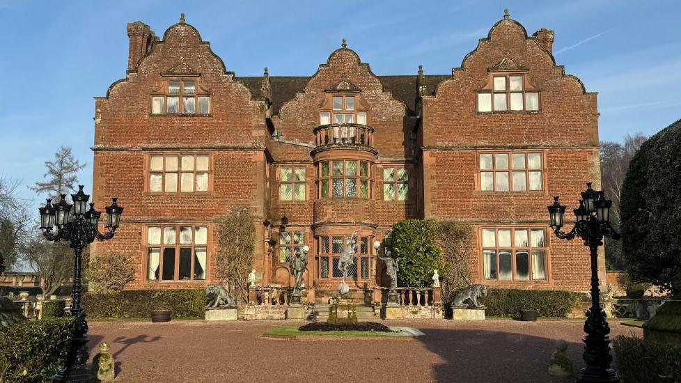 A red-brick mansion with a turning circle and stone lion statues either side of a bay window. Blue sky at the top of the image and trees in the background. 