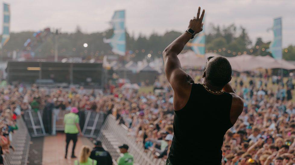 The back view of a performer, with his left arm raised and his head tilted back as he sings, and in front of him crowds, held back with a narrow u-shaped barrier, and in the background more tents and lights and banners