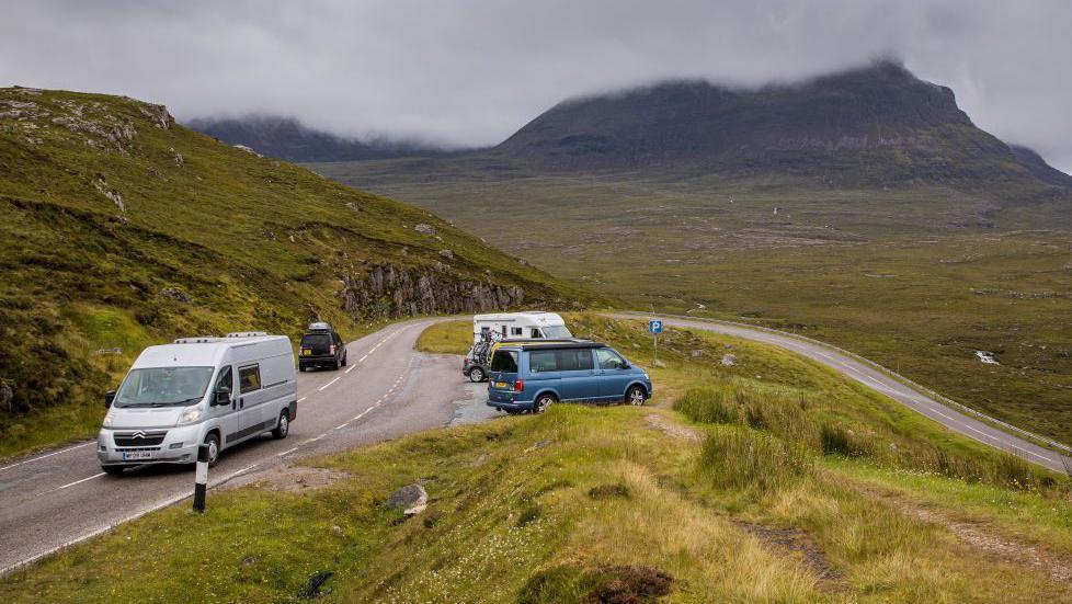 Traffic on a road and parked in a parking area on the NC500 near Ullapool