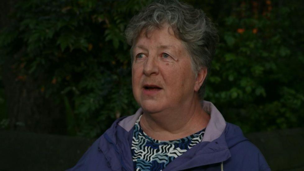 A woman with short grey hair stands in front of a hedge. She is wearing a blue top with a white, grey and black print, and a purple coat.