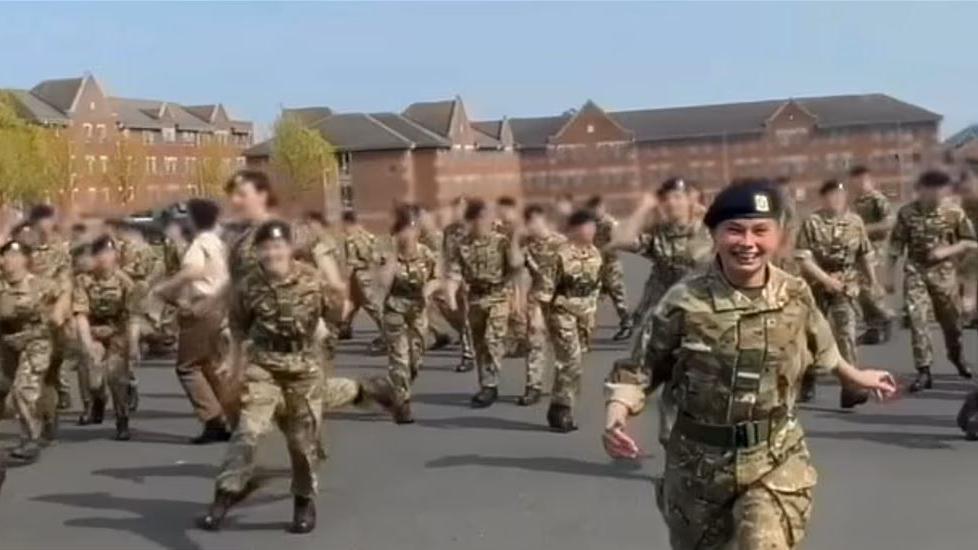 A screenshot from a video showing a regiment practicing their uniform marching and saluting on camp at Larkhill. There is a large group of around 30 people dressed in green and brown camouflage uniforms and black caps. They are standing on a large tarmac area surrounded by fields and trees. In the background there is a brown brick building with lots of windows.