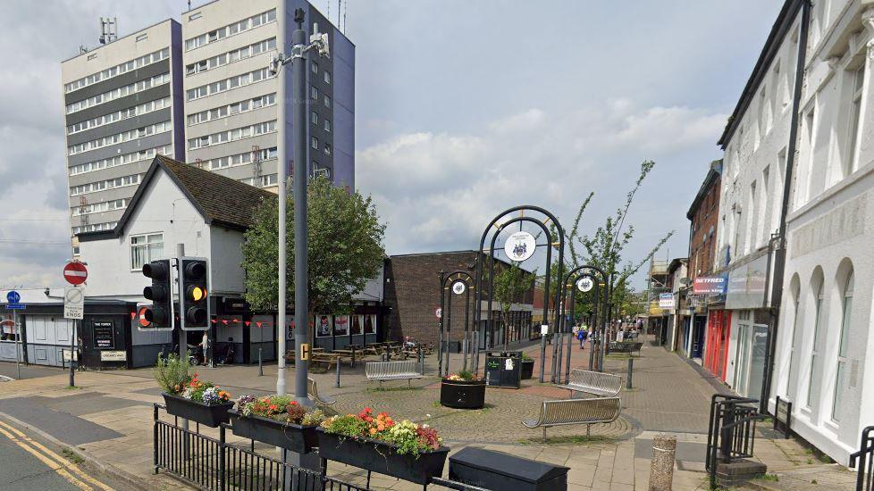 A view looking down Liscard Way high street, a traffic light sits in the foreground, with the Liscard Way metal entrance behind it as one person walks down the empty street with little shops.