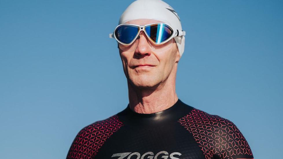 A close up of Martin, he wears a swimming cap and goggles and looks out into the distance, he wears a black wetsuit.