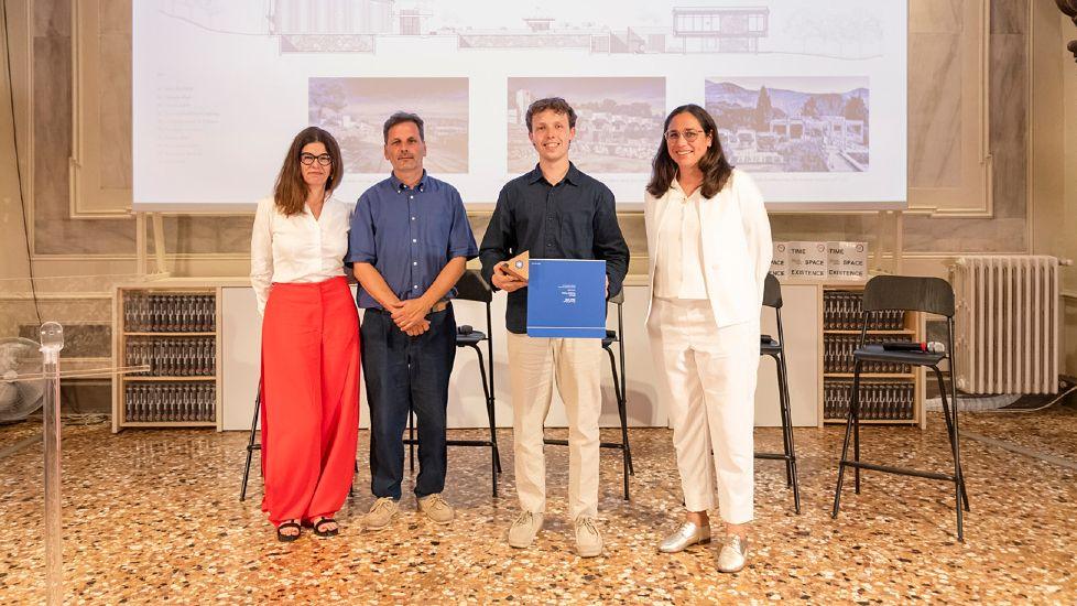 James Langlois holding his award. He is wearing a blue top and cream trousers. To his right a woman, with shoulder length brown hair, wearing all white. To hie left are a man and a woman. The man is in a blue shirt and dark blue trousers. The woman has should length brown hair, is wearing a white top and has red trousers. The floor is marble. Behind each of them are plastic chairs.