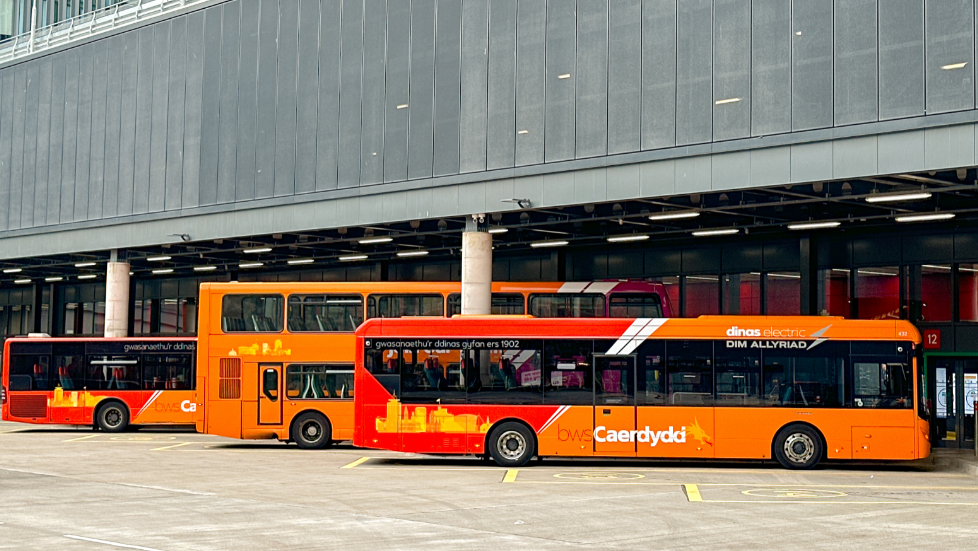 A picture of Cardiff Bus interchange by Cardiff Central Railway station. There are three buses parked in bays outside. They are orange in colour. In front of them is a large glass wall, with pillars interspersed.