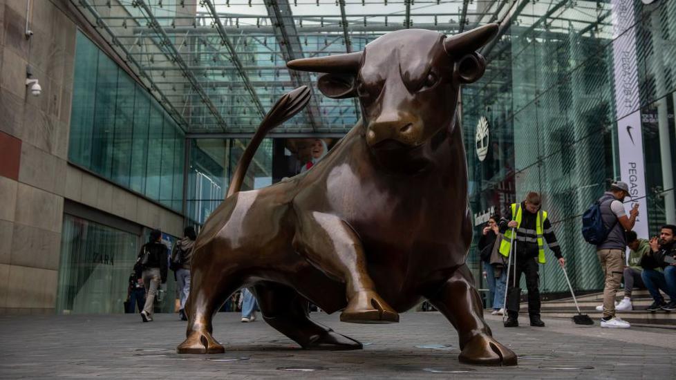 A large bronze statue of a bull outside a shopping centre, which has a glazed front. The bull has one leg raised and an angry expression, as if it is about to charge. Pedestrians and a street cleaner in a fluorescent jacket can be seen behind it.