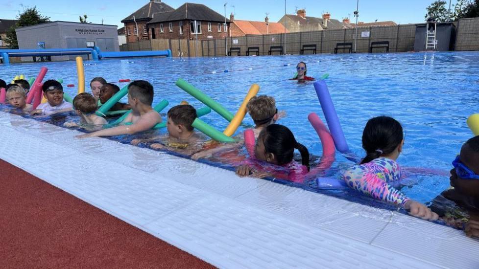 Several pupils with woggles holding on to the side of the pool with a woman in goggles in the middle 