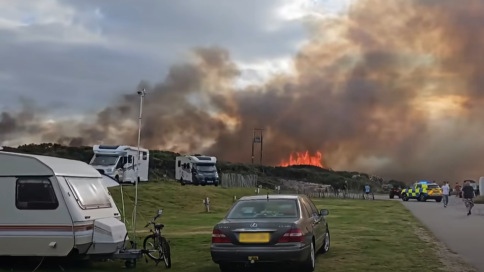 Orange flames leaping above grassland next to a campsite. There are two campervans and a caravan on the site, along with people and cars. There is also  an emergency vehicle