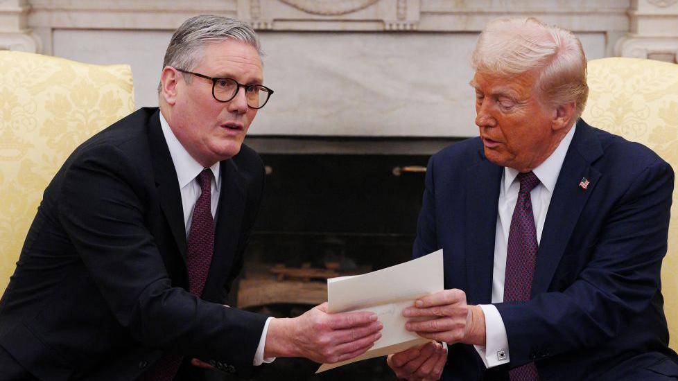 Prime Minister Sir Keir Starmer in the Oval Office presenting a letter to Donald Trump. Trump is wearing a blue suit, white shirt and purple tie. Starmer is wearing a black suit, white shirt and purple tie.