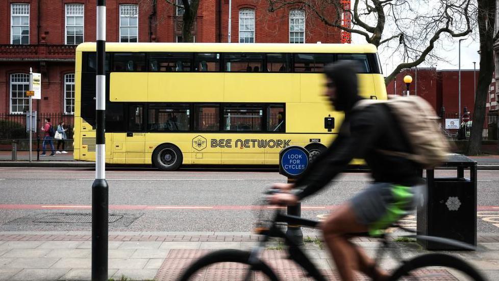 A yellow double-decker bus with 'Bee Network' written on the side stands at a bus stop. A man on a bicycle rides past in the foreground.