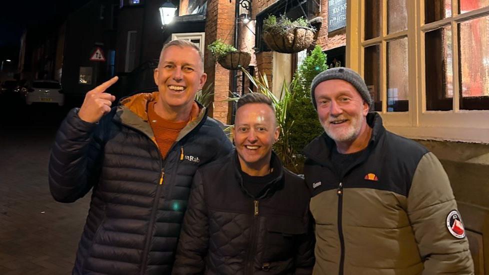 Three smiling men in a street at night, one has grey hair and a blue jacket and is pointing upwards, another is shorter, with a black jacket and dark hair and the third has a grey beard and grey hat and is wearing a grey and black coat