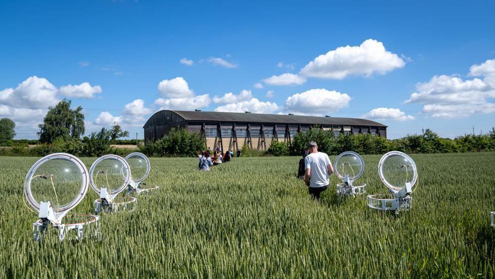 A field of green crops with a farm building in the background, and several students walk through the field. There are five circular pieces of equipment which appear to be monitoring some of the crops.