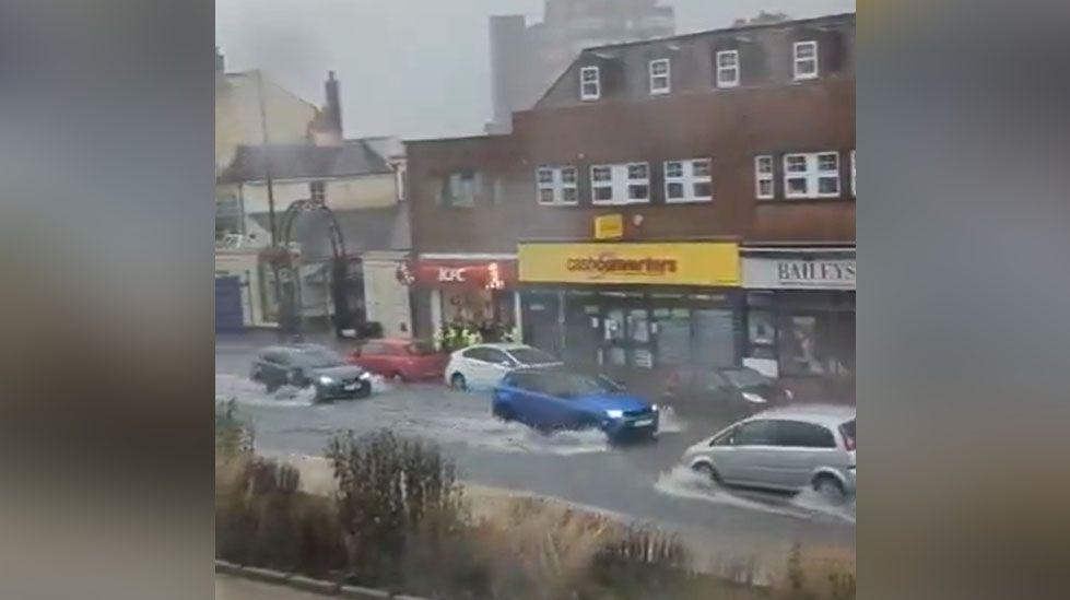 Cars driving in floodwater in front of shops on Dunstable High Street.