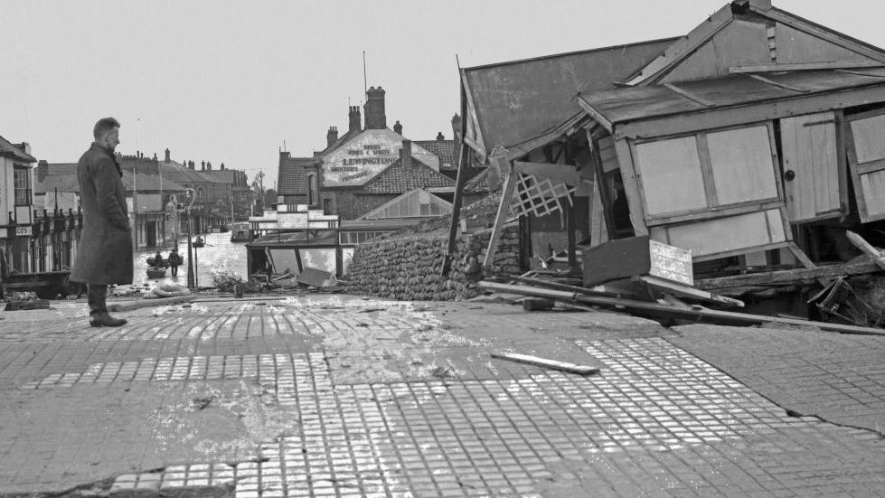 Archive black and white image showing the devastation caused by flooding in Mablethorpe in 1953. A man is standing looking at a wooden building which has collapsed.