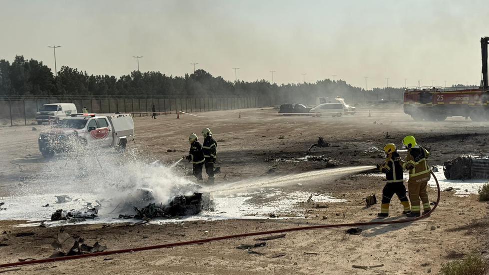Firefighters work at the scene of a plane crash, water spraying onto a burnt piece of metal on the ground, it is in an open stretch of land with cars and a fence visible in the background.