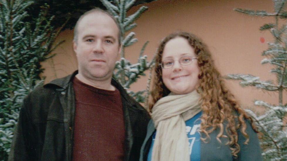 A man with brown hair wearing a maroon jumper and black denim jacket stands next to a woman with brown long curly hair wearing a blue shirt and cream scarf. They are smiling and stood in front of some Christmas trees with snow on the branches.