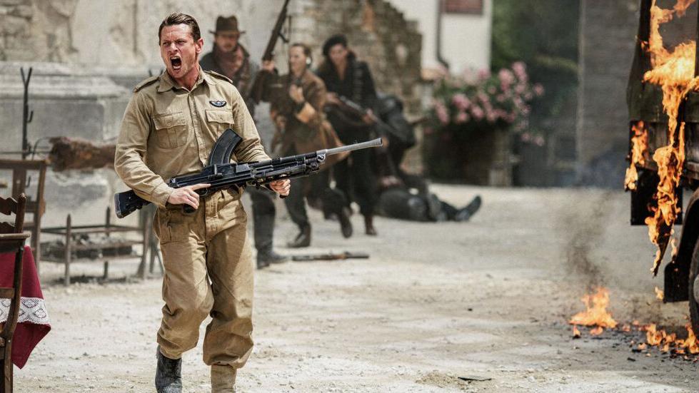 Jack O'Connell, playing Blair Mayne in Rogue Heroes. He is wearing a khaki military uniform and holding a black rifle. He is shouting as a lorry burns to the right-hand side of the screen.