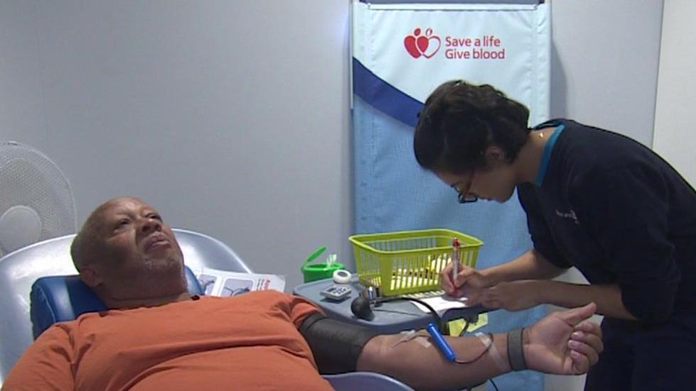 Isaac is seen lying back in a blood donor chair while a health worker takes notes next to him.