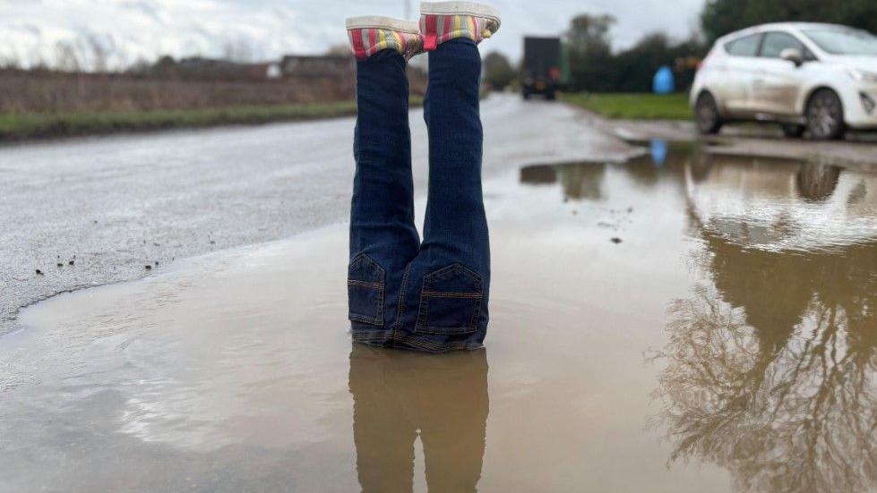 A pair of fake legs are placed in a water-filled pothole with the feet sticking out at the top. The legs are constructed from blue jeans and there are colourful shoes on the fake feet. The pothole is large and is at the side of a road. The water is muddy. There is a white car in the background and a green lorry further down the road.