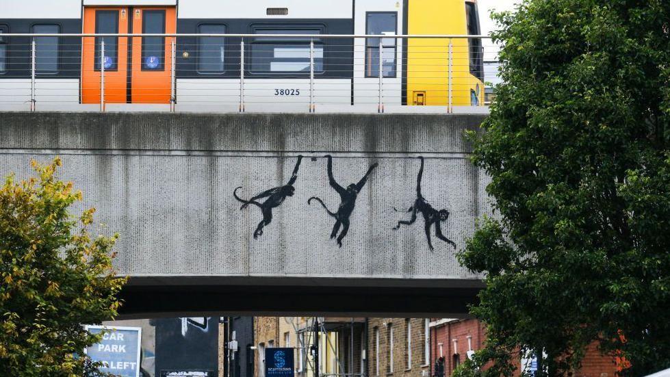 Three black monkeys appearing to hang from a grey concrete railway bridge in Brick Lane