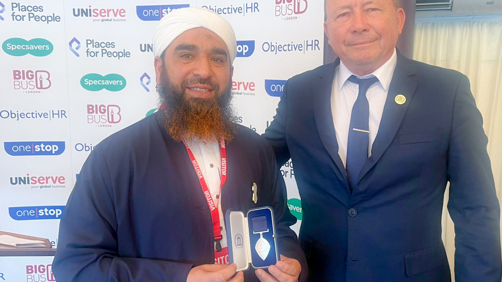 Syed Muhammad Faisal Sami holding his award at the ceremony. He has a white turban, a blue top over a white shirt and a long dark beard. He holds a small case with a pointed metal badge attached to a ribbon in it. Next to him is a white man in a blue suit