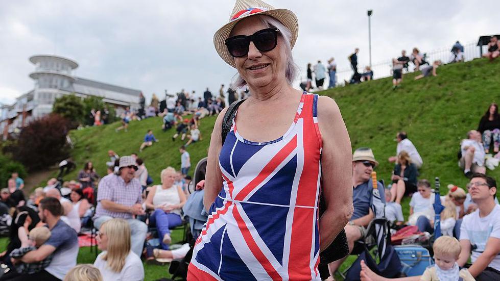 Woman dressed in a red, white and blue Union flag dress looking into a camera at an Armed Forces Day event in Cleethorpes. She is also wearing sunglasses and a hat with crowds of people sat enjoying the event in the background.