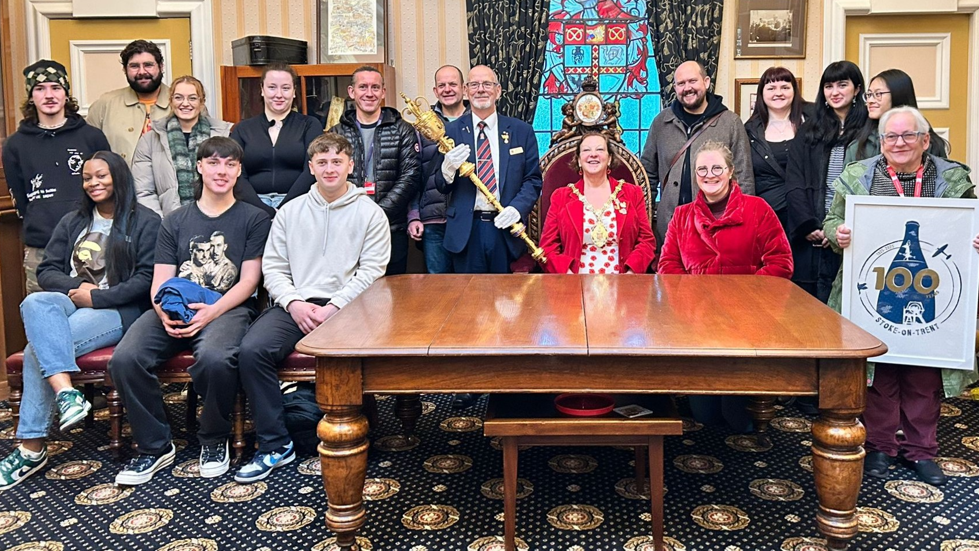 A gathering of people, including a man carrying a mace and a woman in a chain of office, sit behind a large wooden table, with one of them holding a picture of a blue triangle with the number 100 on it