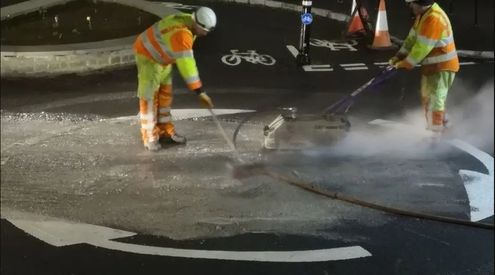 Men in high-vis jackets and trousers use machinery to resurface a roundabout. The roundabout is a hollow circle formed by white arrows